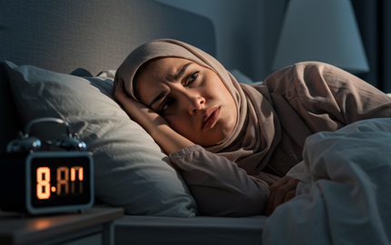 A stressed woman in a hijab lies awake in bed, looking anxiously at the time.
